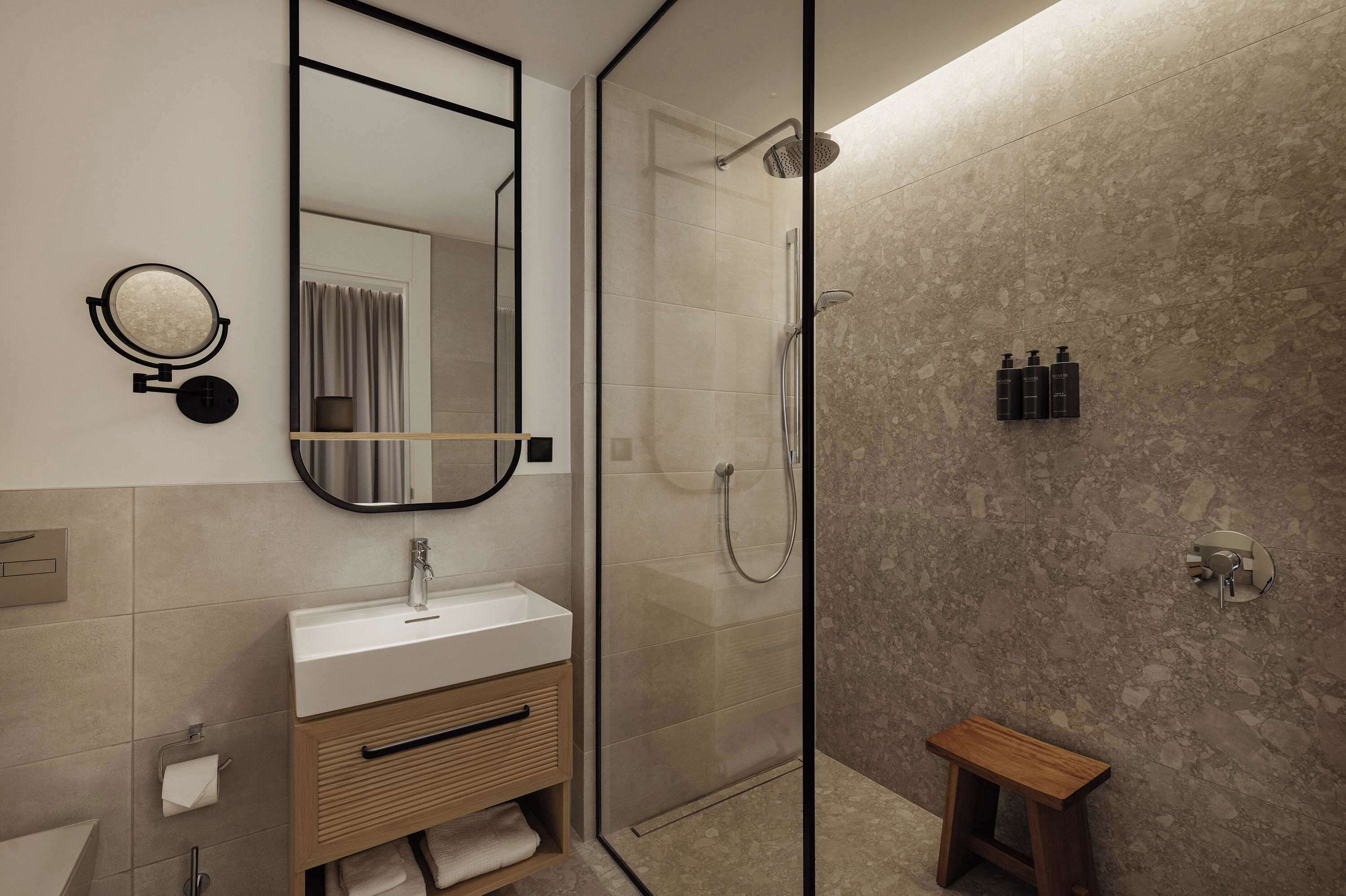 A modern bathroom with a glass-enclosed shower, a large mirror, a wooden vanity, and a small stool.