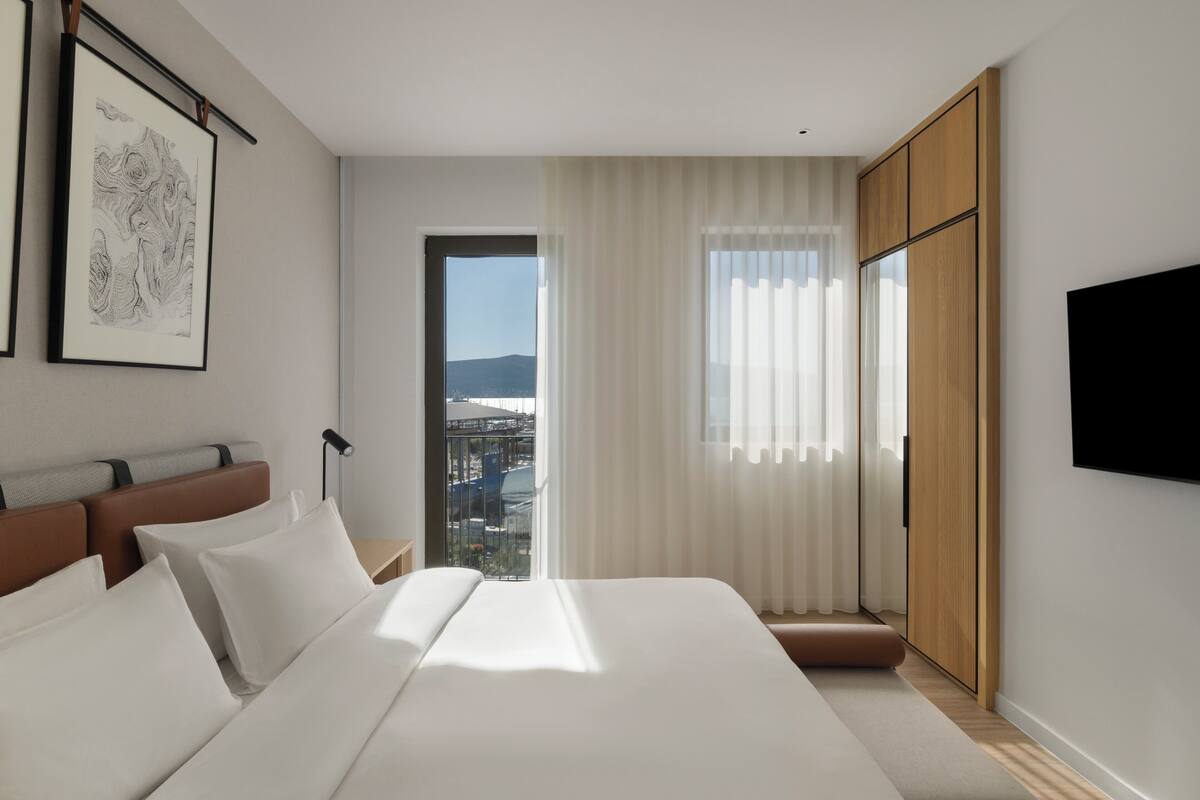 A modern hotel room with a large bed, a view of the city, and a mounted TV.