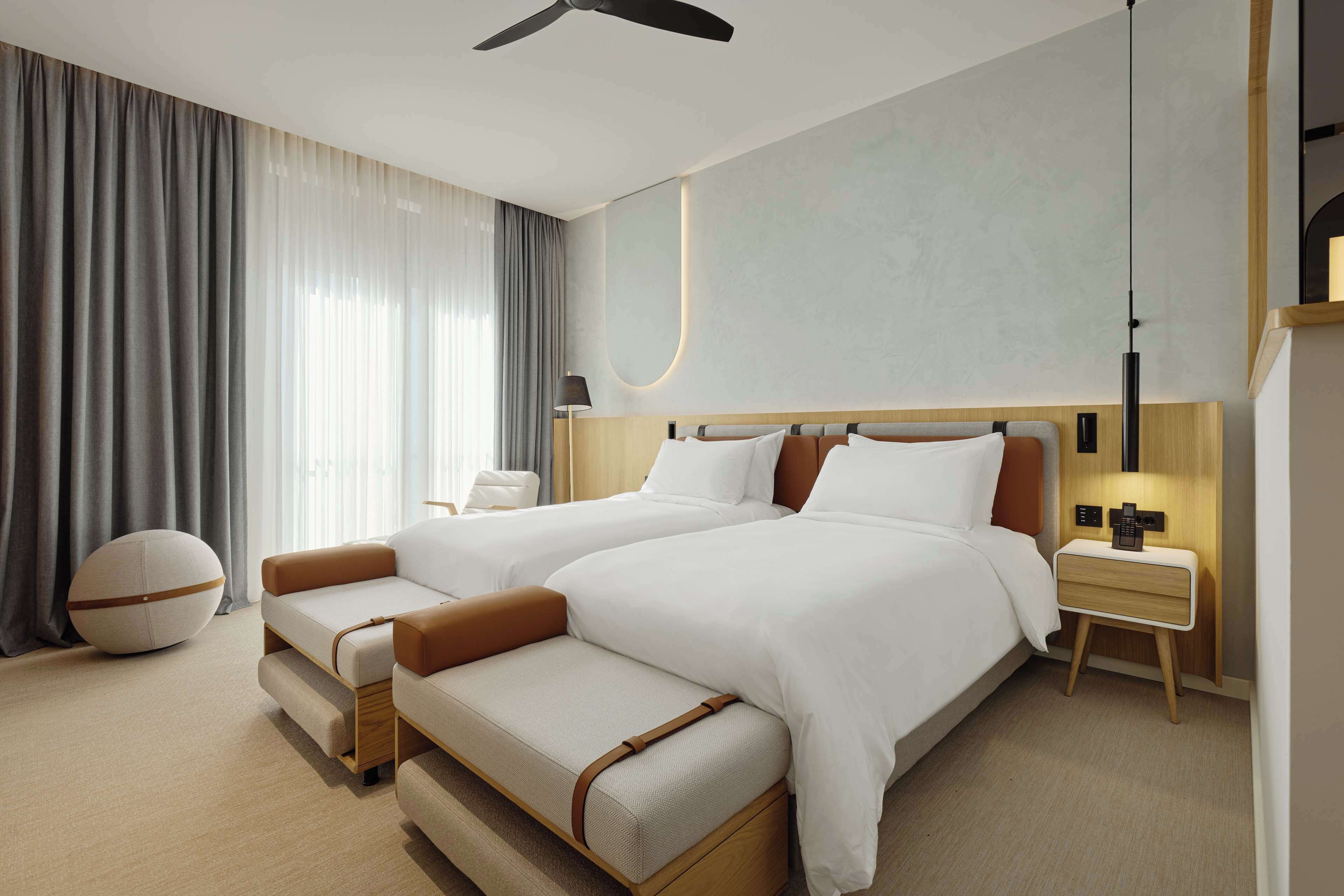 A hotel room with two beds, a round ottoman, a nightstand with a lamp, and a ceiling fan.