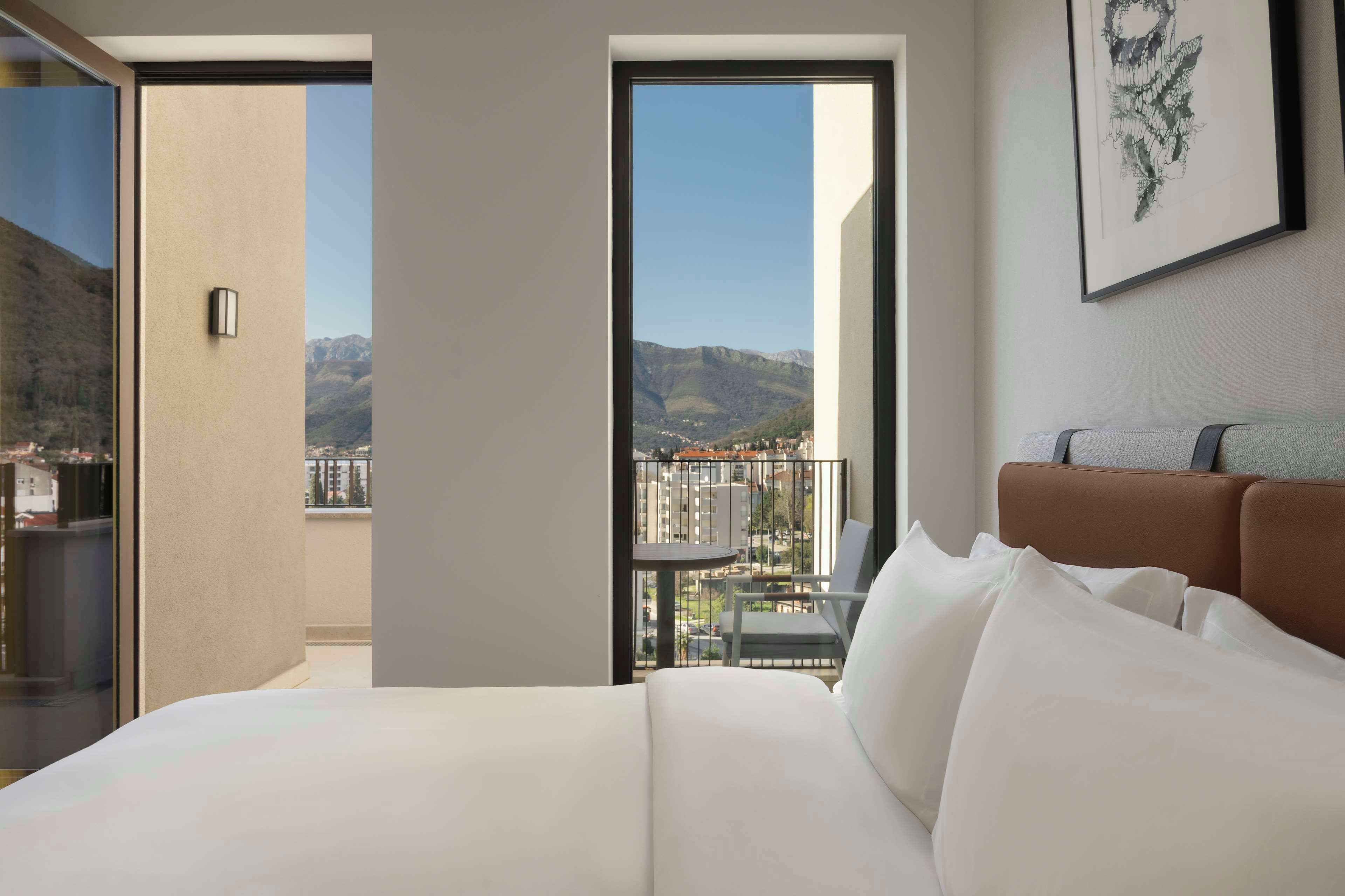Habitación de hotel con cama, una silla, un balcón y vistas a la ciudad y las montañas.