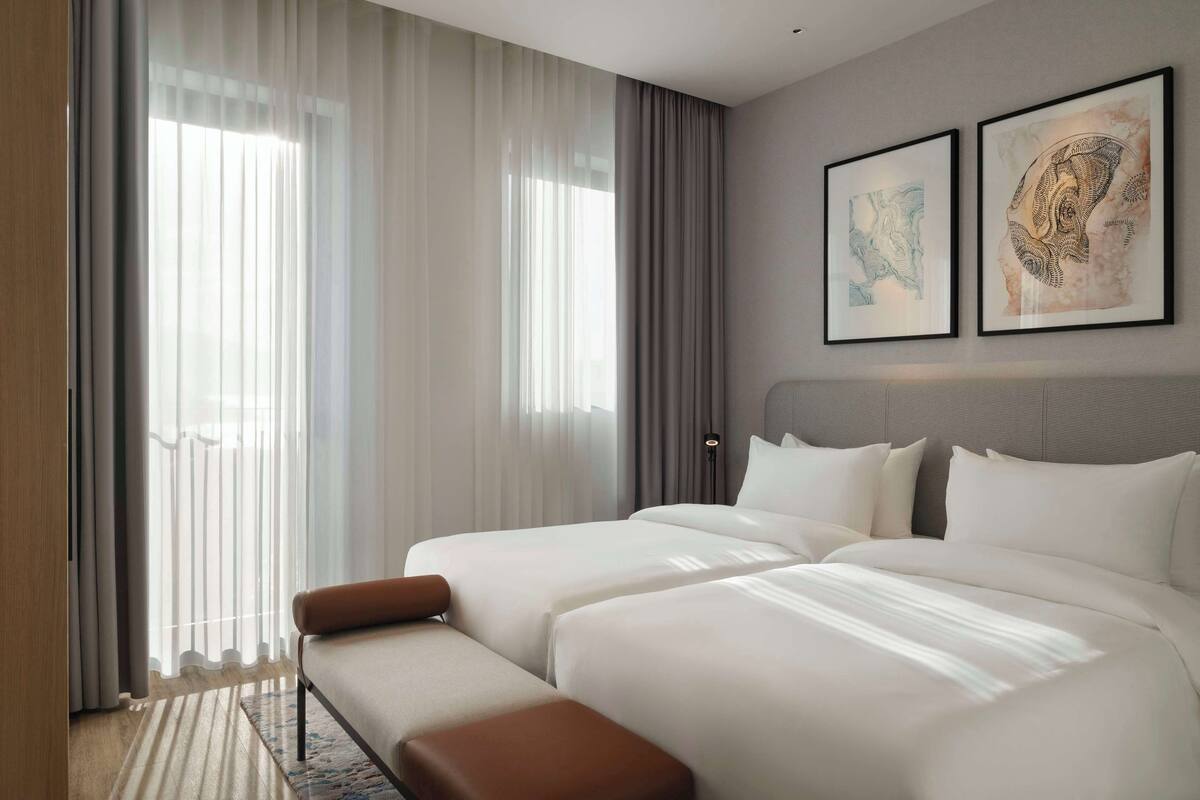 A hotel room with two beds, a bench, a large window with sheer curtains, and two framed artworks on the wall.