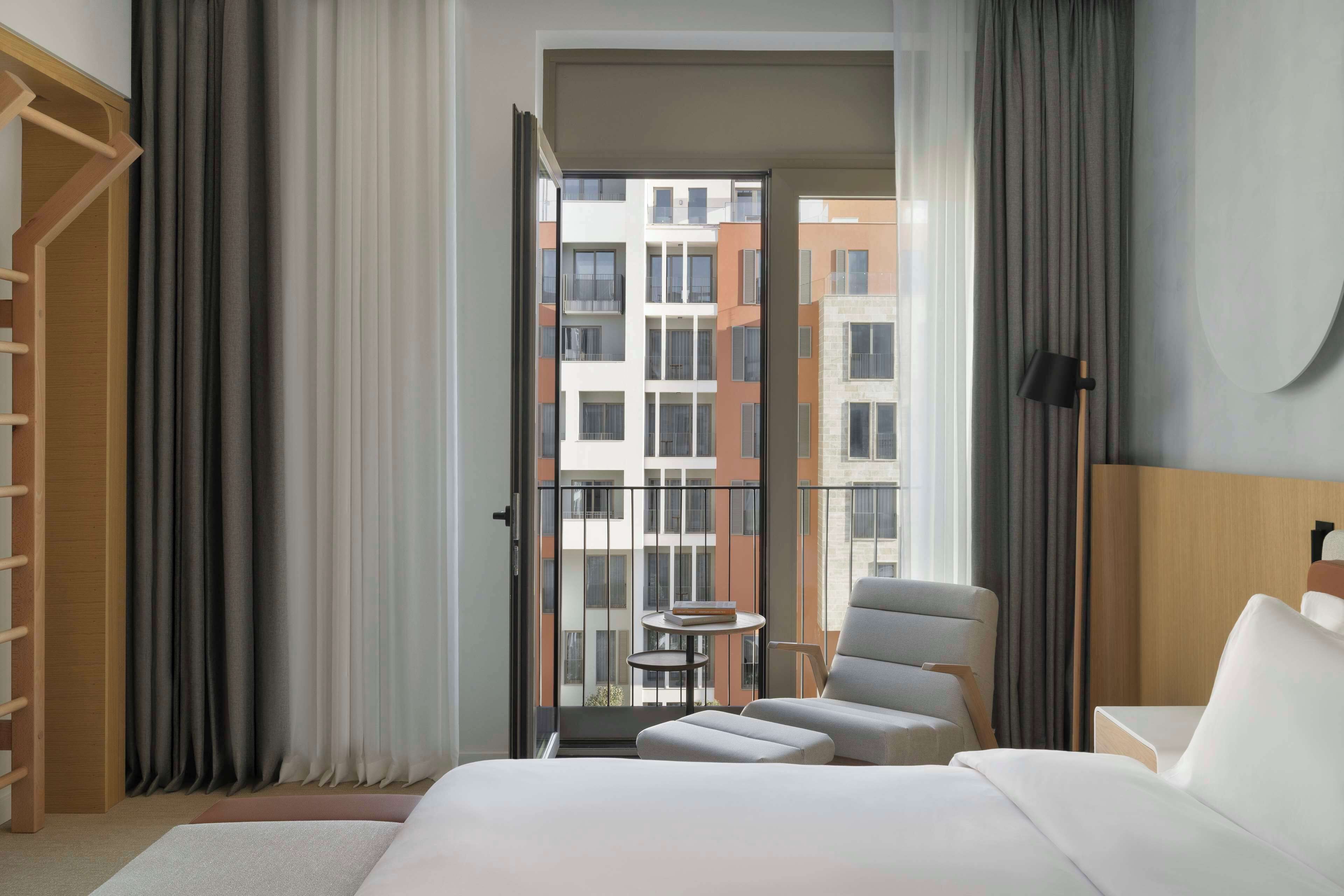 A hotel room with a bed, a chair, a small table, and a view of a building through a glass door.