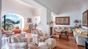 A spacious living room with a large window, a fireplace, and a variety of seating options. - Villa Amelia in Capri (Capri)