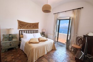 A bedroom with a bed, a nightstand, a chair, a television, and a view of the sea through a large window.