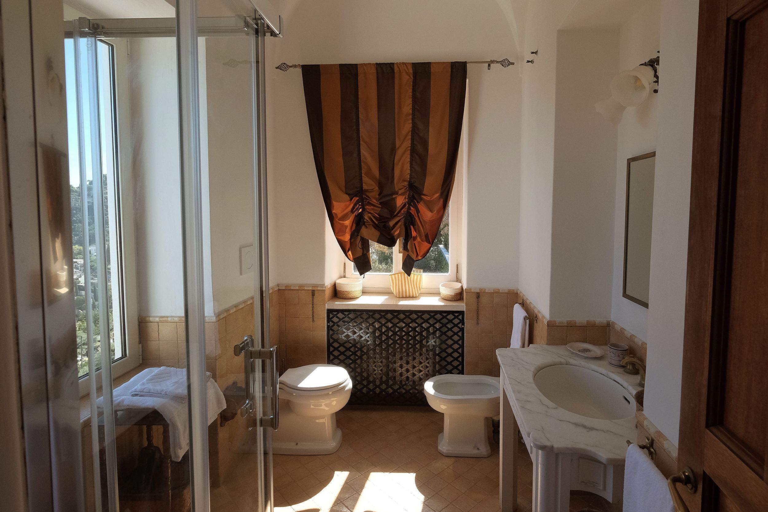 A bathroom with two toilets, a shower, a sink, and a window with curtains.
