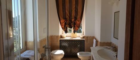 A bathroom with two toilets, a shower, a sink, and a window with curtains.