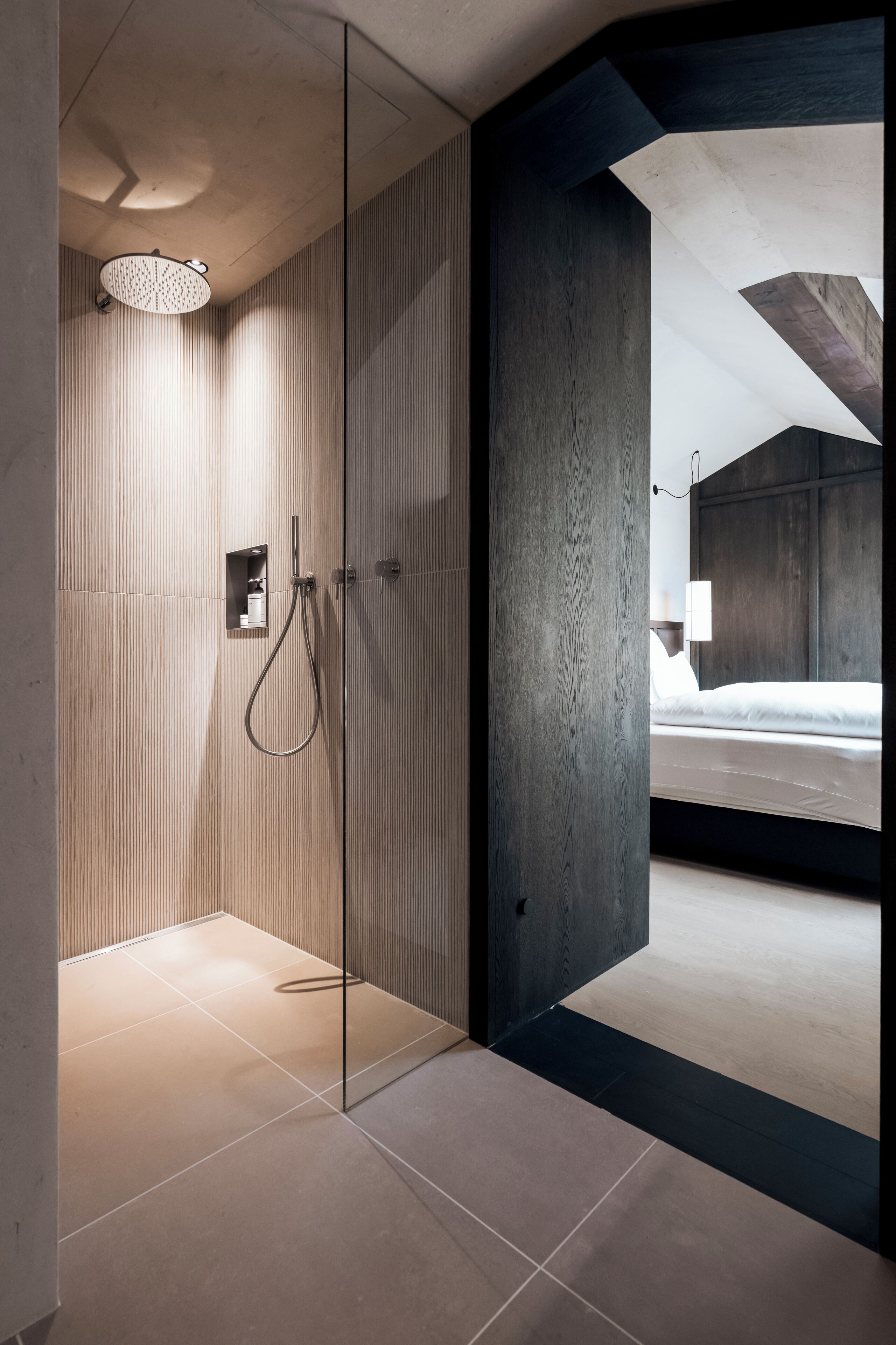 Un baño moderno con cabina de ducha de vidrio, un panel de pared de madera y una habitación visible con cama y mesita de noche.