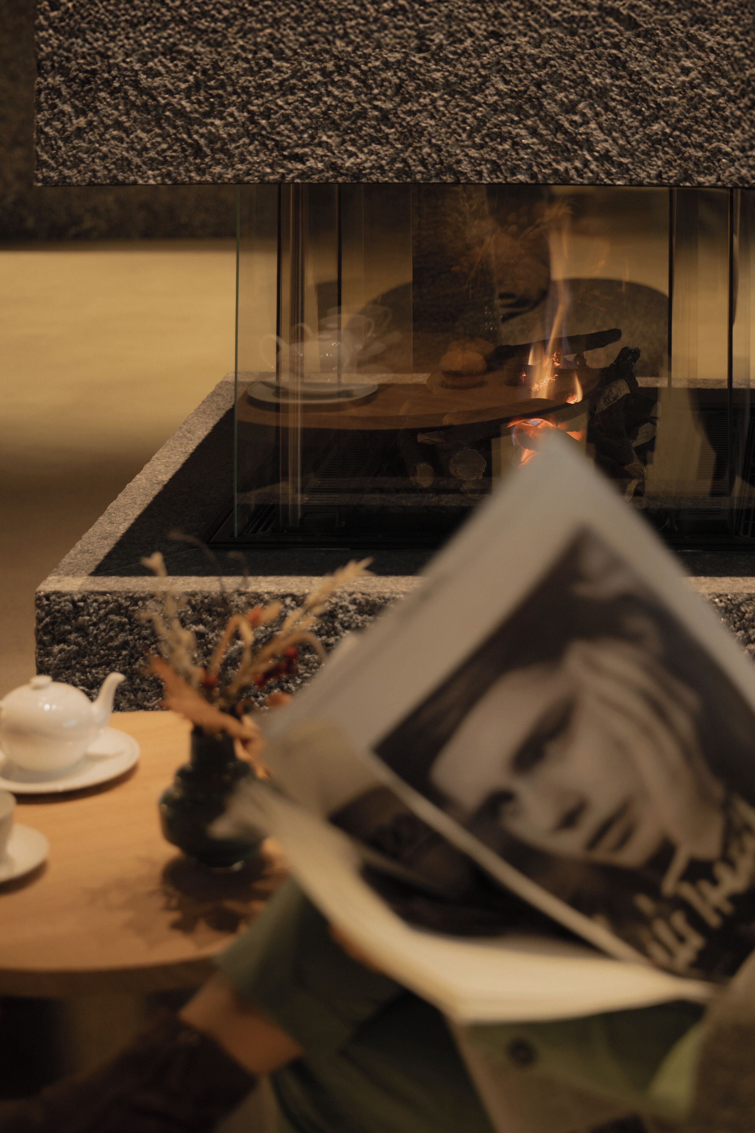 A room with a fireplace, a table with a teapot and a vase of dried flowers, and a person reading a magazine.