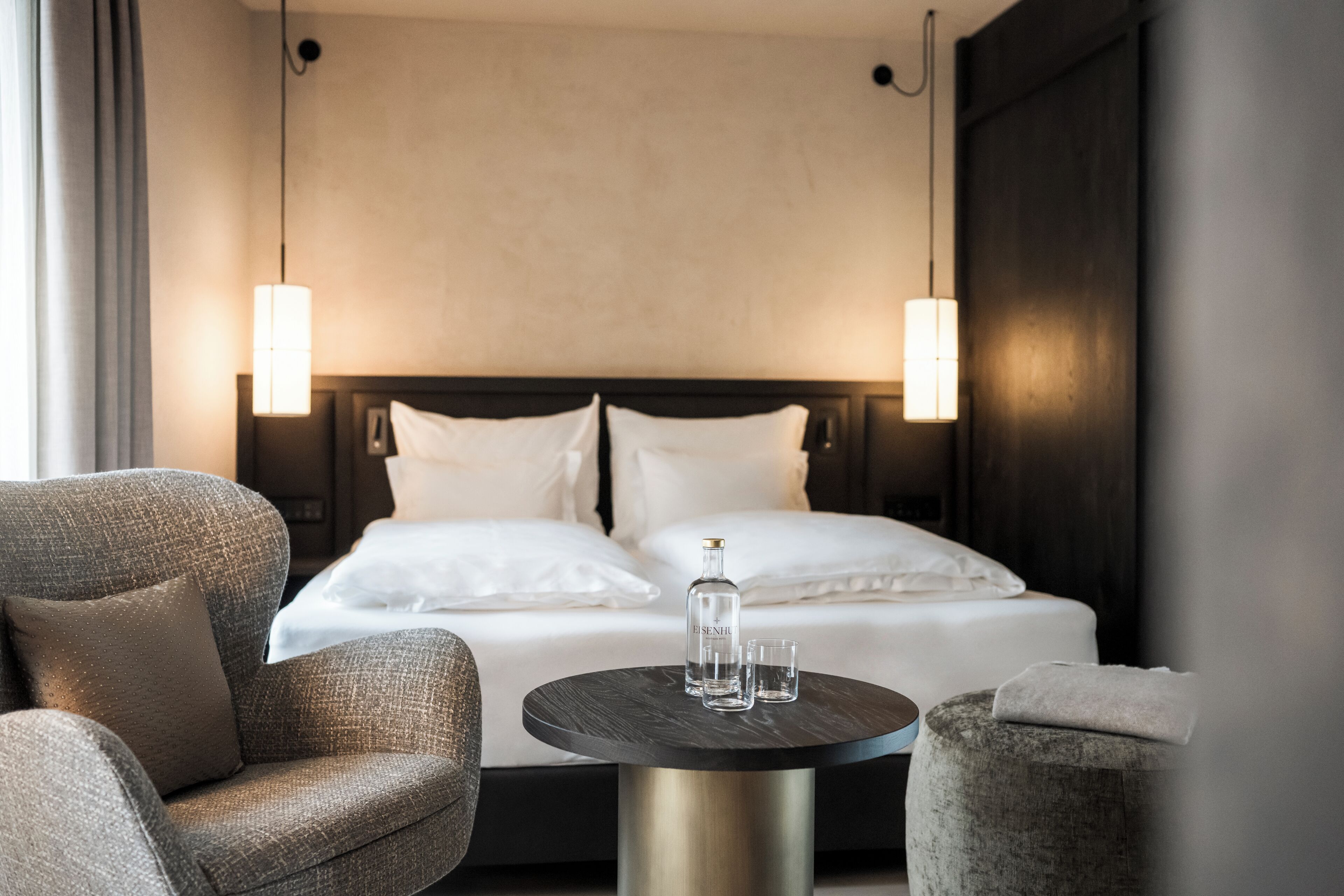 A hotel room with a bed, two bedside lamps, a round table with a bottle and glasses, and two armchairs.