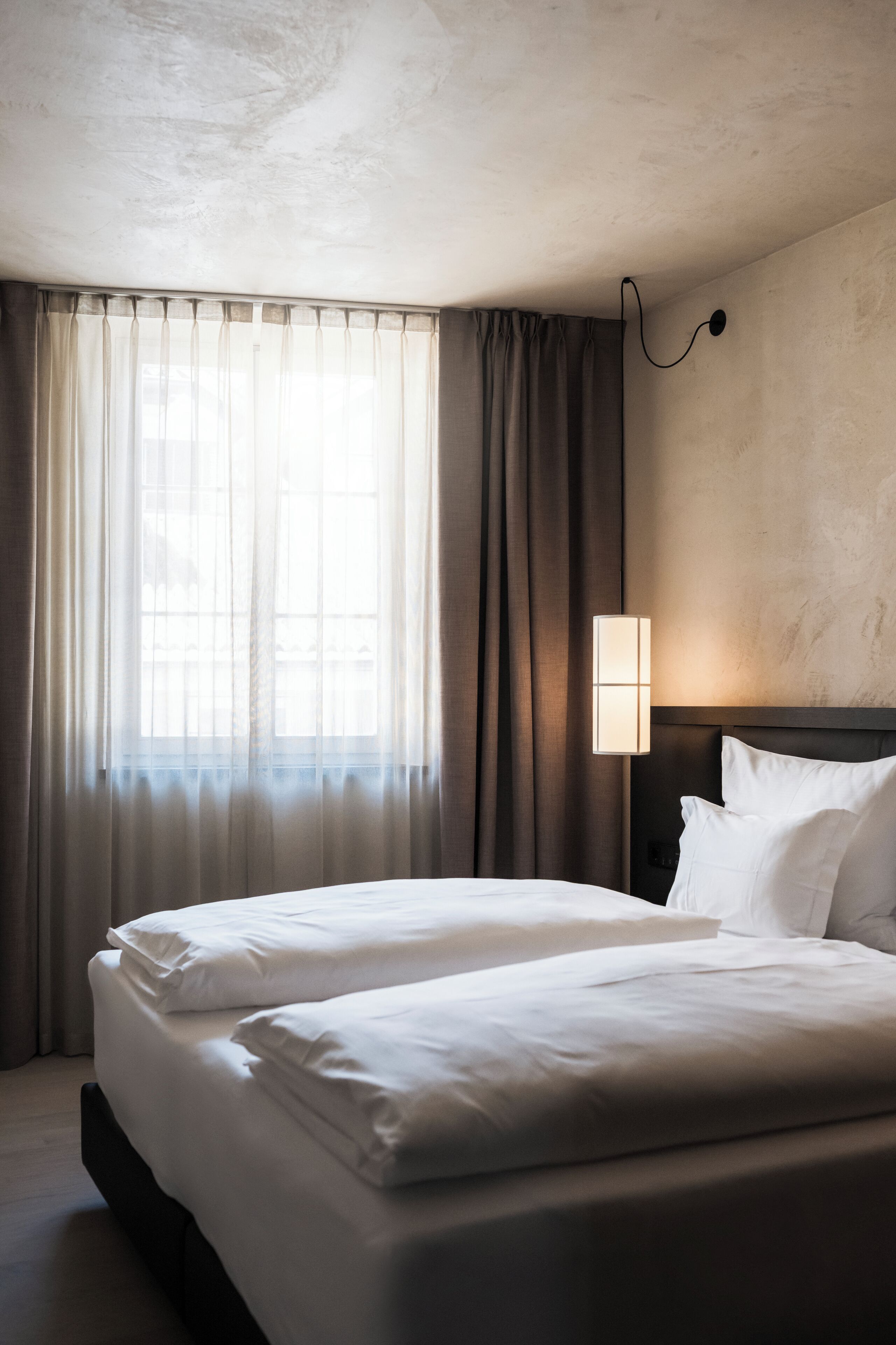 Camera d'albergo con due letti, un'ampia finestra con tende leggere e una lampada a parete.