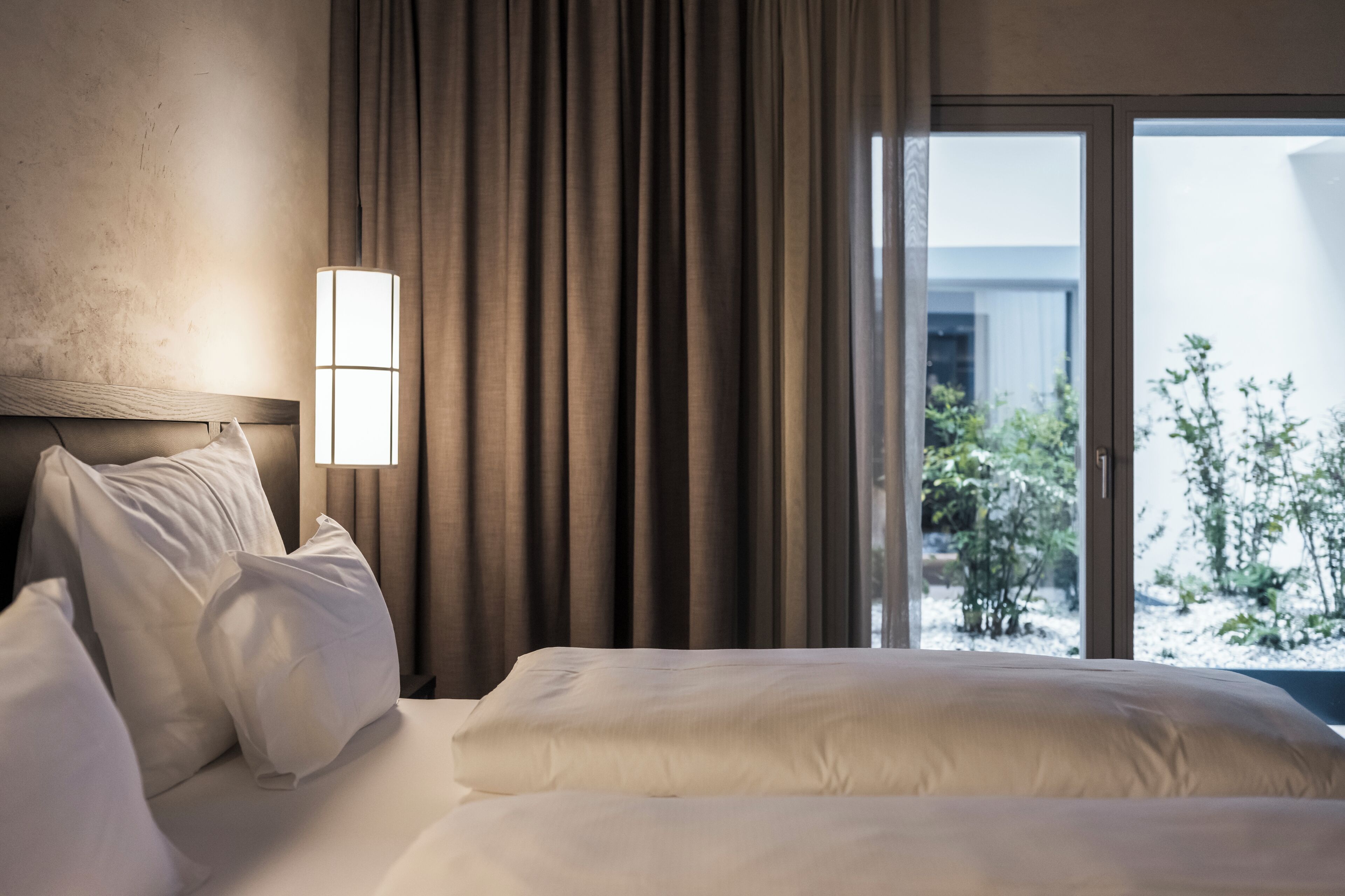 Habitación de hotel con cama, mesita de noche, lámpara y vista al exterior a través de un ventanal grande.