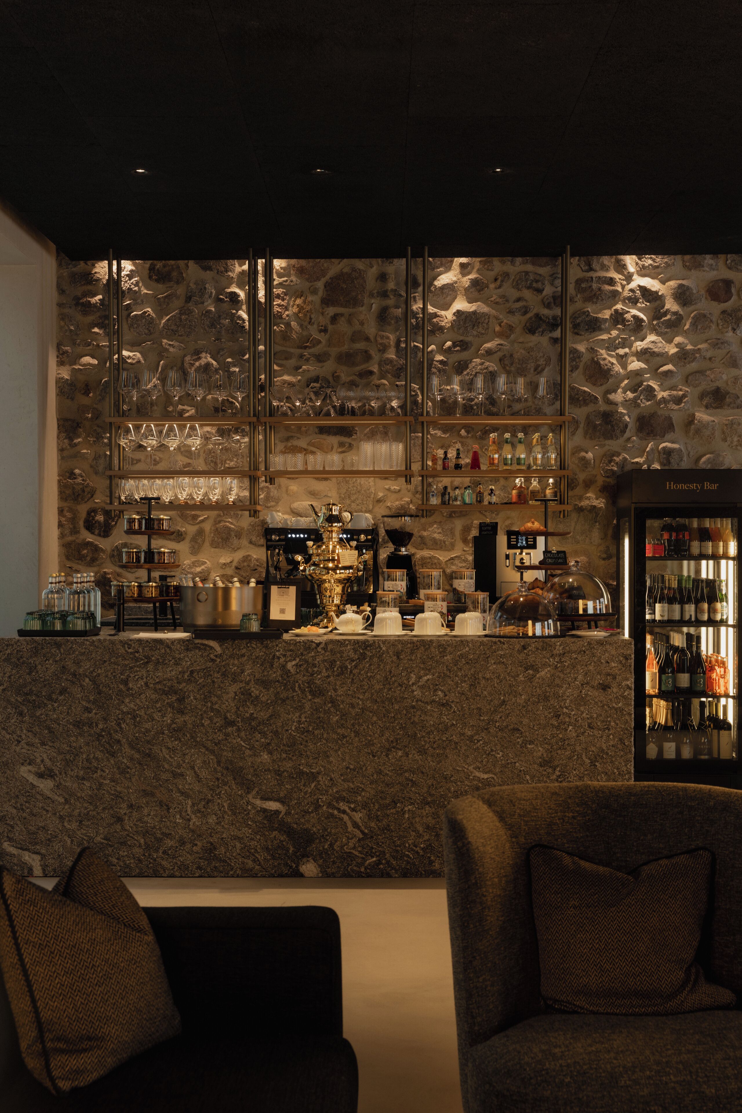 A bar area with a stone wall, shelves with glasses and bottles, and a coffee machine. There are two chairs in the foreground.