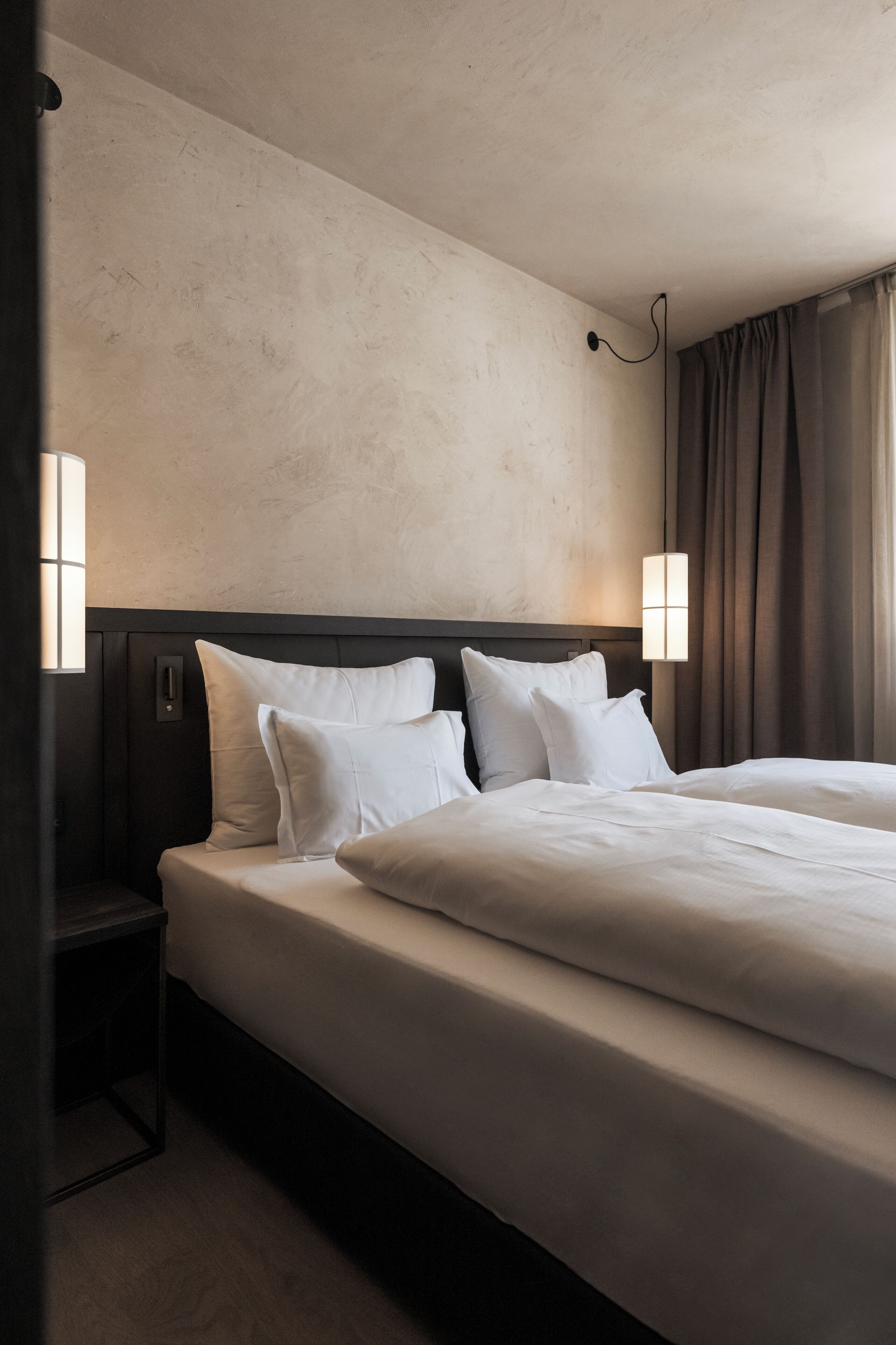 A hotel room with a large bed, two bedside tables, a wall-mounted lamp, and a window with curtains.
