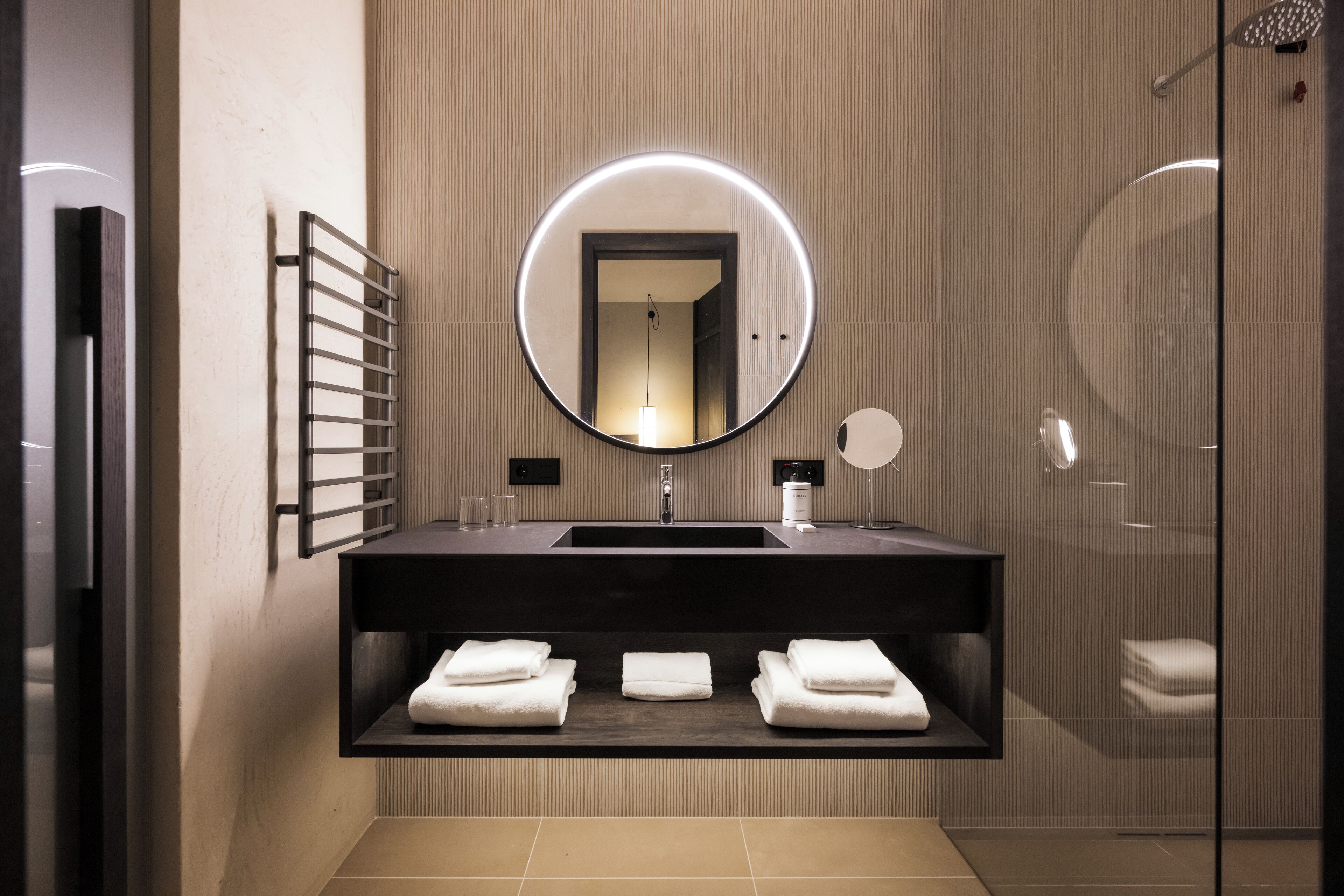 Un baño moderno con un espejo redondo, un lavabo oscuro y tres toallas dobladas ordenadamente en un estante.