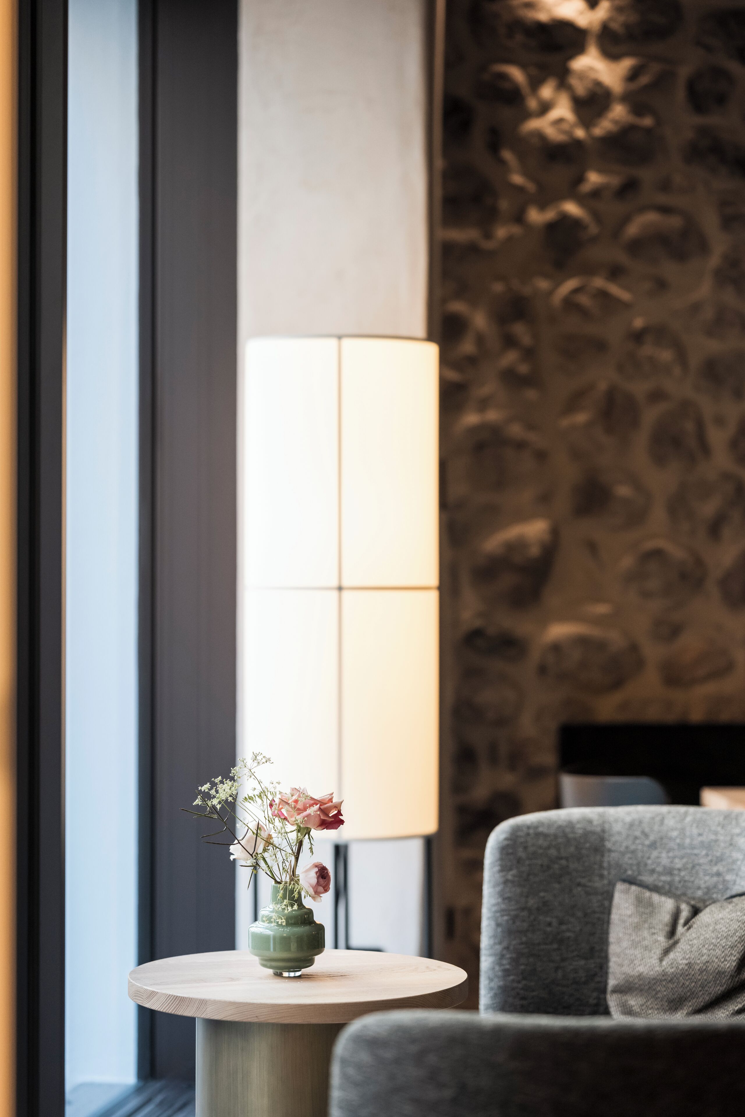 Una habitación moderna con una mesa redonda, un florero con flores, una lámpara y un sillón gris.