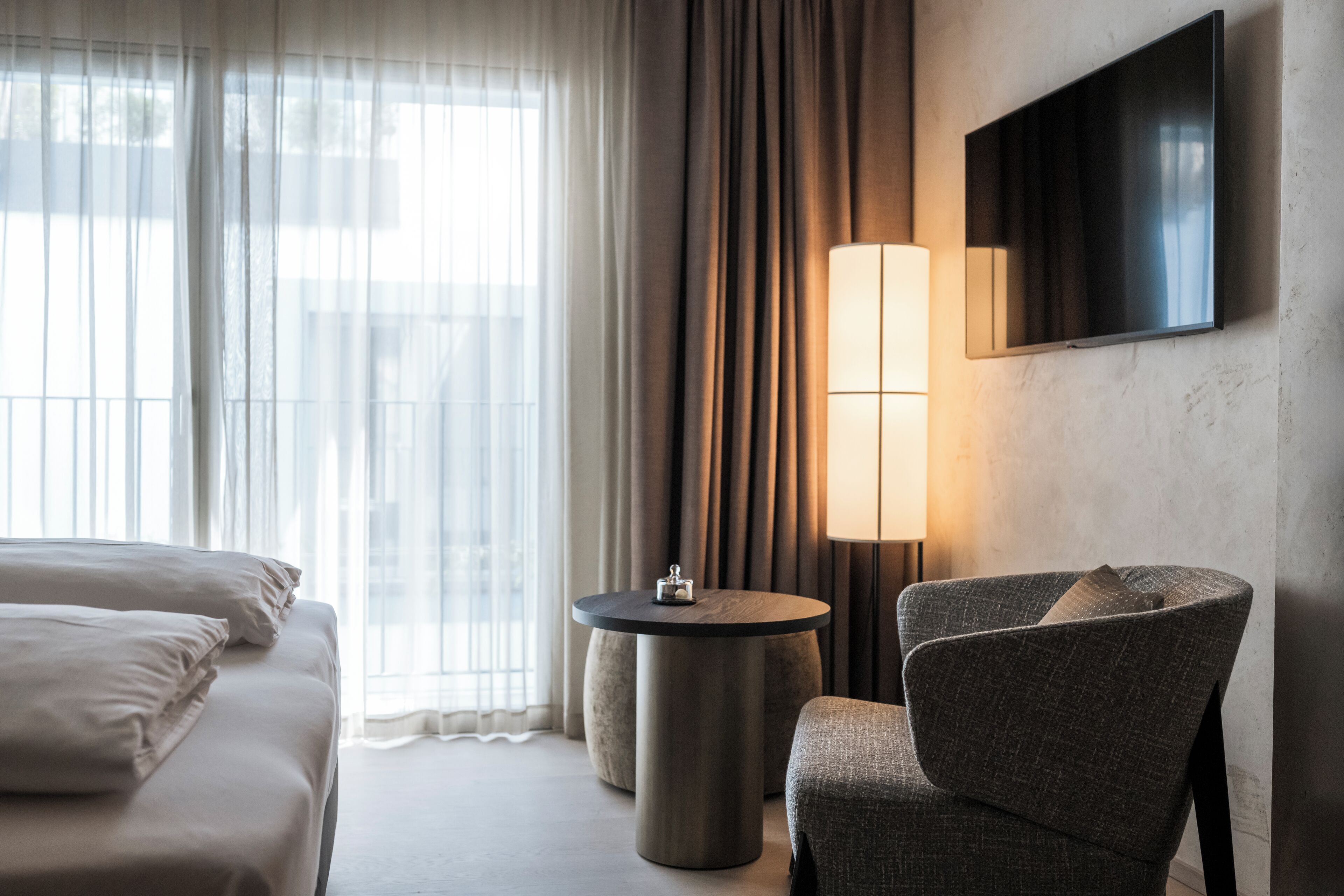 Camera d'albergo con un letto, un tavolino rotondo, una sedia, una TV a parete e una finestra con tende.