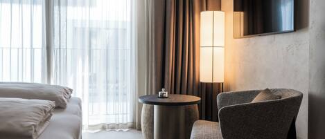 A hotel room with a bed, a round table, a chair, a TV mounted on the wall, and a window with curtains.