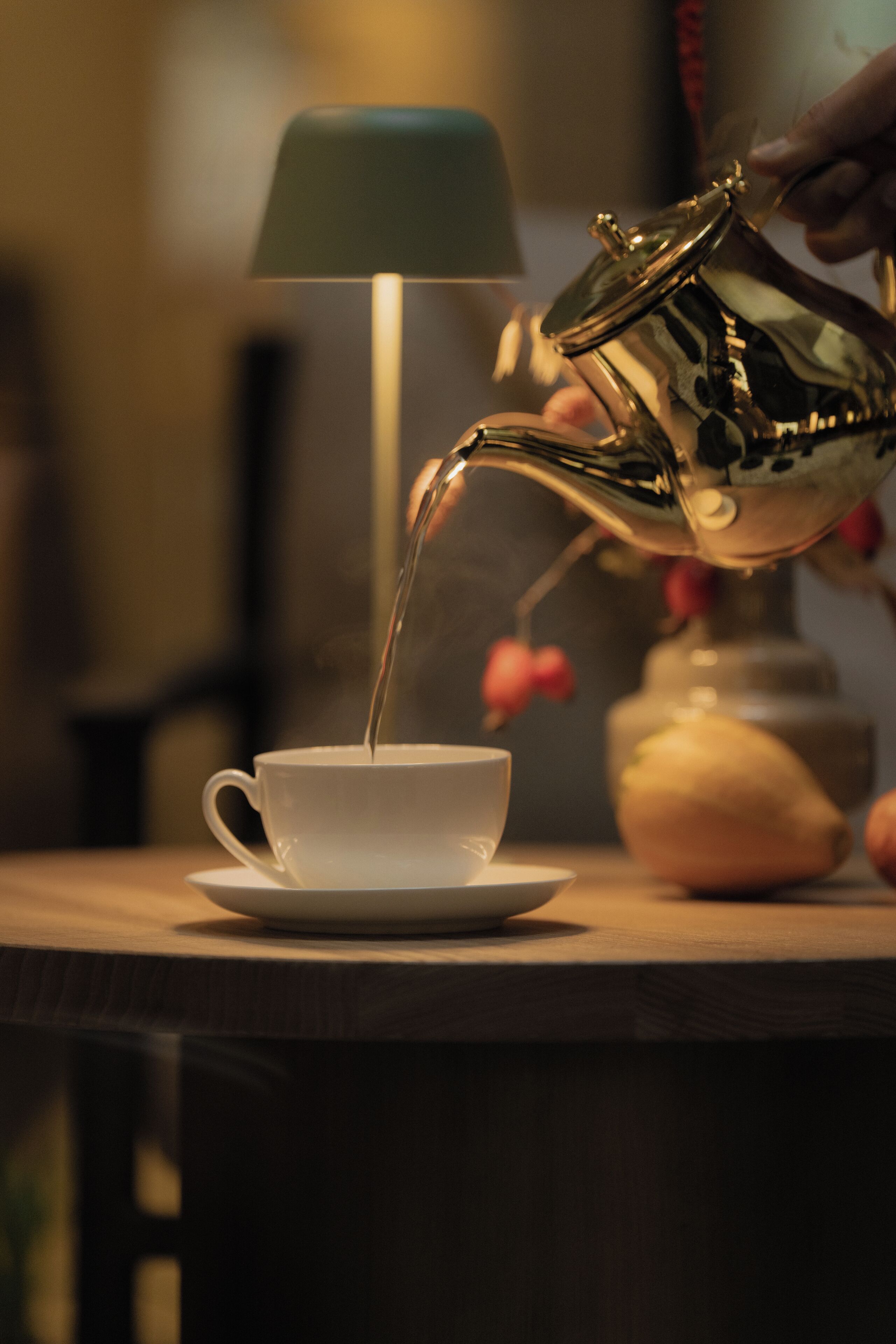 A hand pouring liquid from a metallic pitcher into a white cup on a saucer.