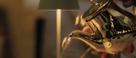 A hand pouring liquid from a metallic pitcher into a white cup on a saucer.