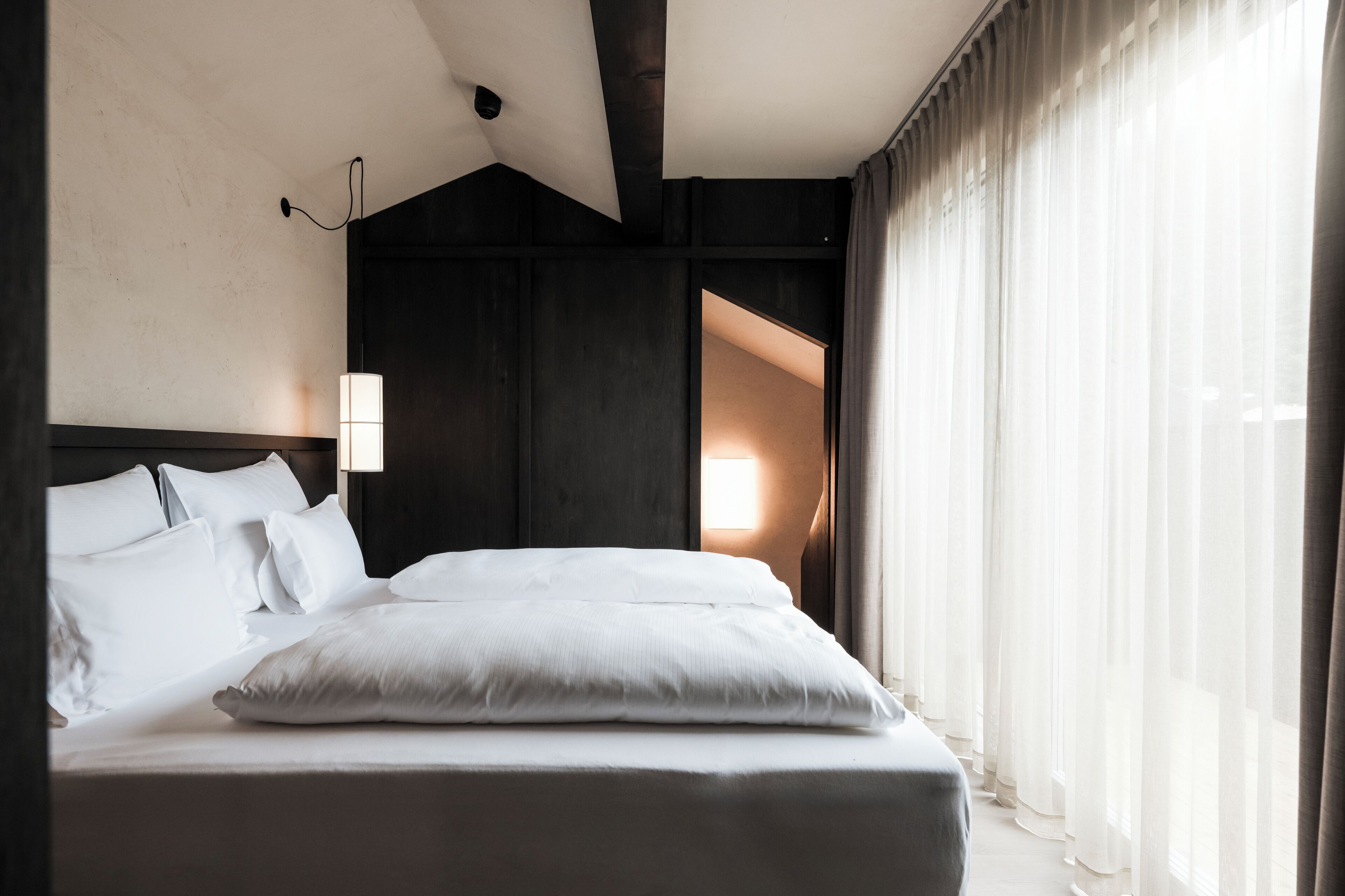 A modern hotel room with a large bed, a bedside lamp, and a view of a bright room through sheer curtains.