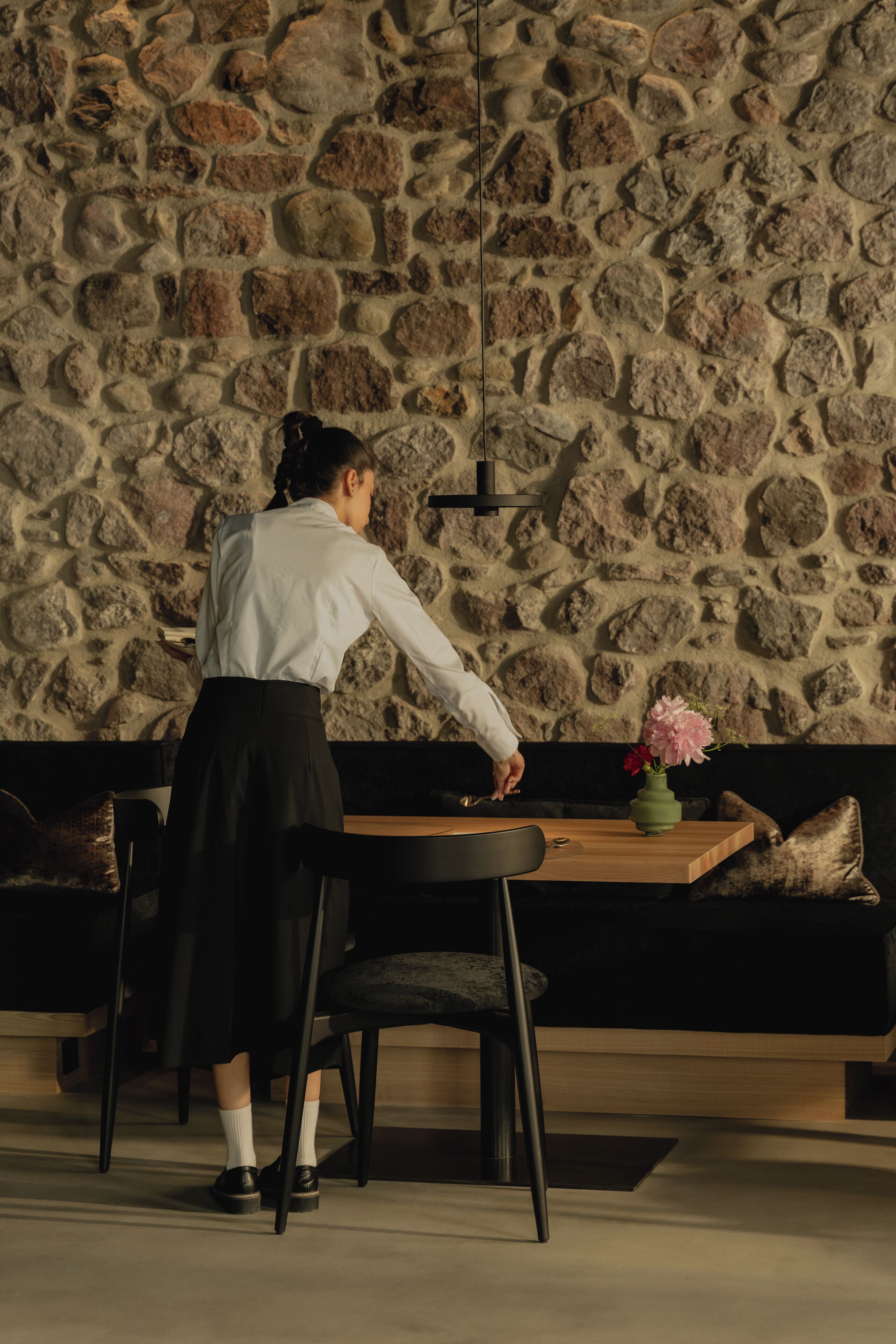 A person standing by a table with a vase of flowers, in a room with a stone wall and a black sofa.