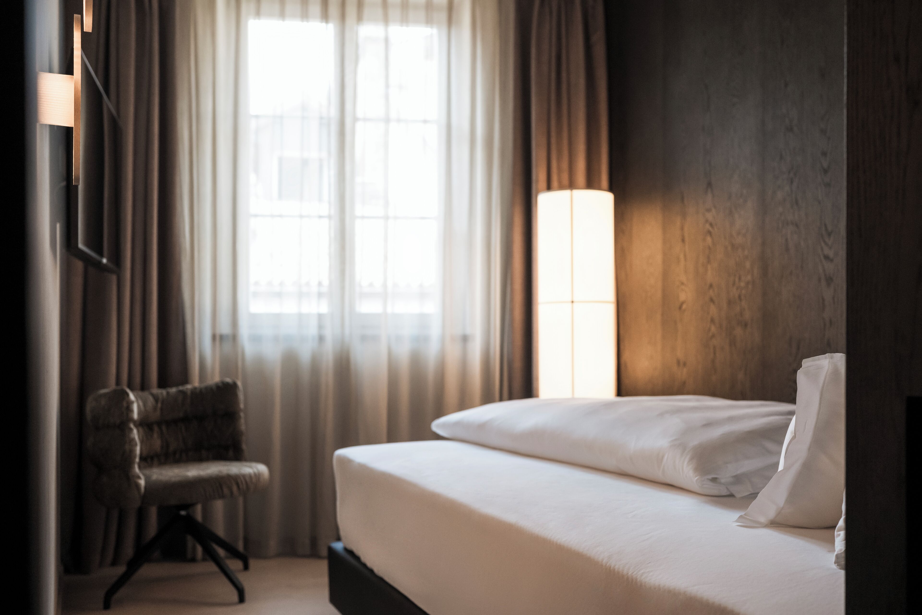 A hotel room with a bed, a chair, a window with curtains, and a wall-mounted lamp.