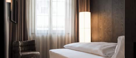 A hotel room with a bed, a chair, a window with curtains, and a wall-mounted lamp.