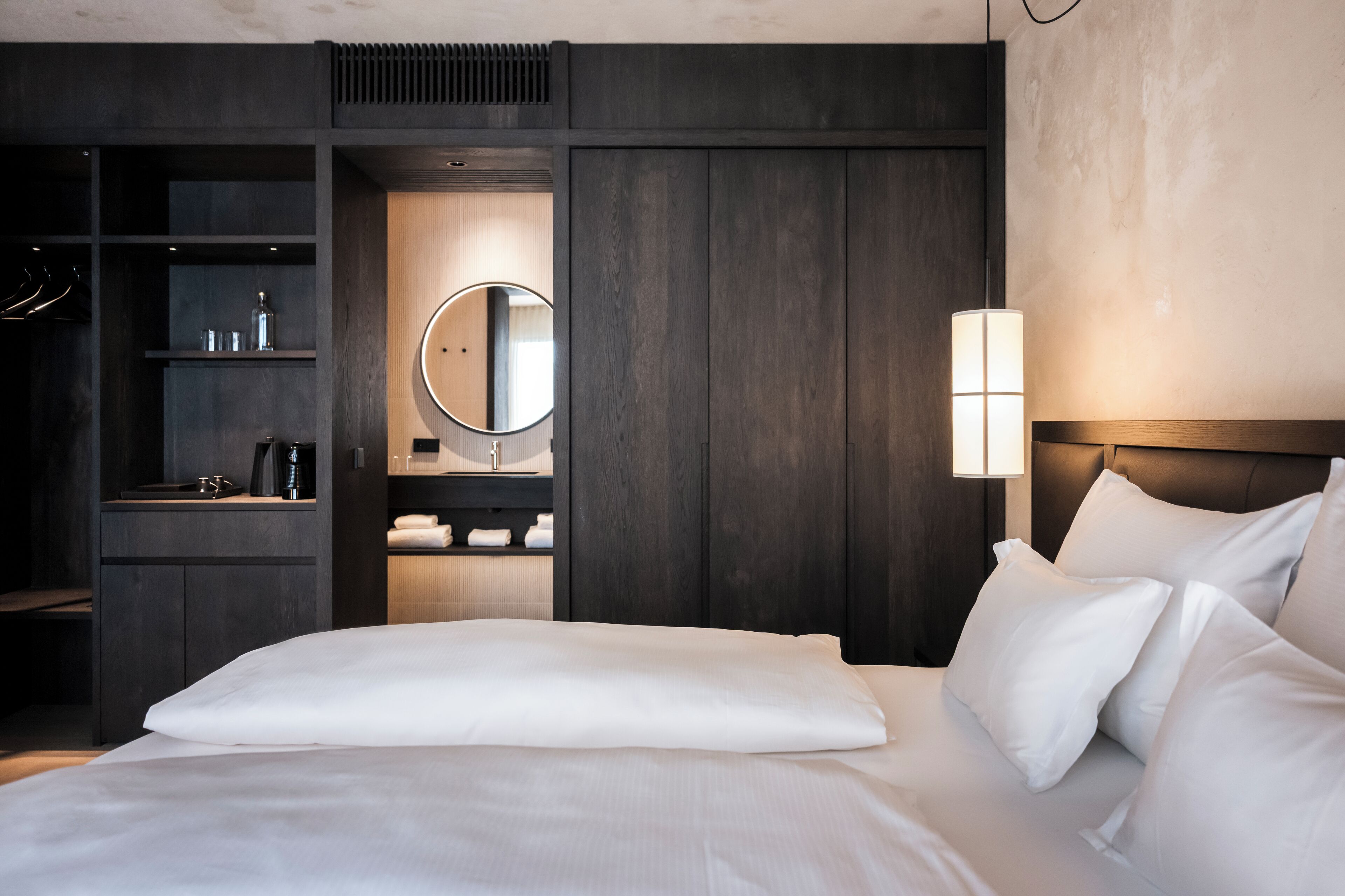 A modern hotel room with a large bed, a built-in wardrobe, a vanity with a mirror, and a bedside lamp.