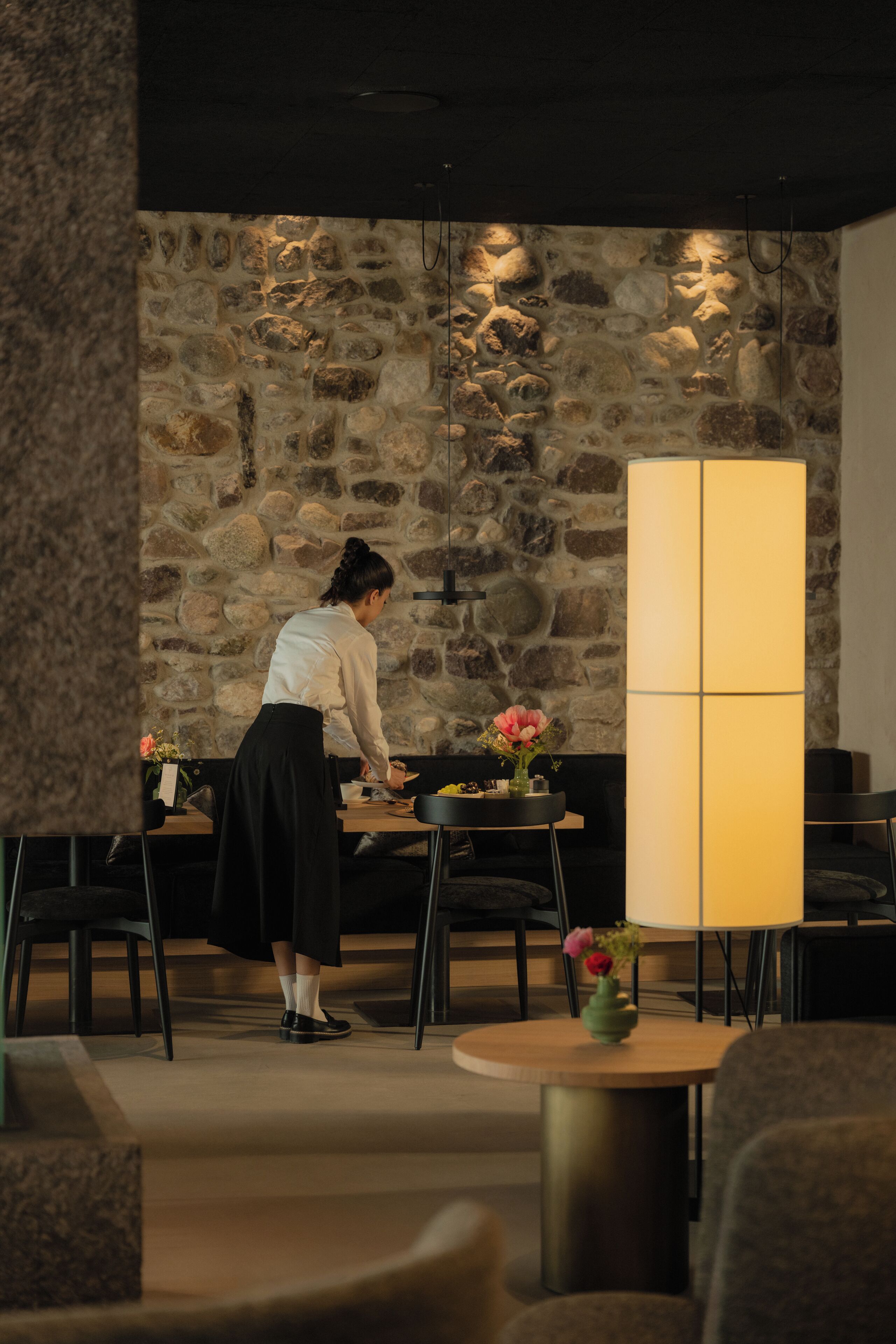 Una persona organizando objetos sobre una mesa en una habitación con pared de piedra y un panel alto, iluminado.