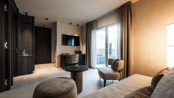 A modern hotel room with a large window, a flat-screen TV mounted on the wall, a round coffee table, a sofa, and a dark wooden wardrobe.