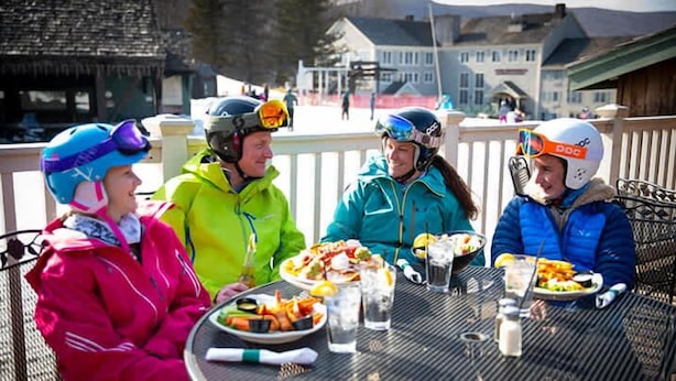 Quatre personnes sont assises à une table, devant de la nourriture et des boissons, et portent des casques de sports d’hiver.