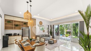A modern kitchen with built-in shelves, a dining area with a wooden table and chairs, and a large sliding glass door leading to a balcony with outdoor furniture. - Kana Village Villa by Kozystay (Tabanan)