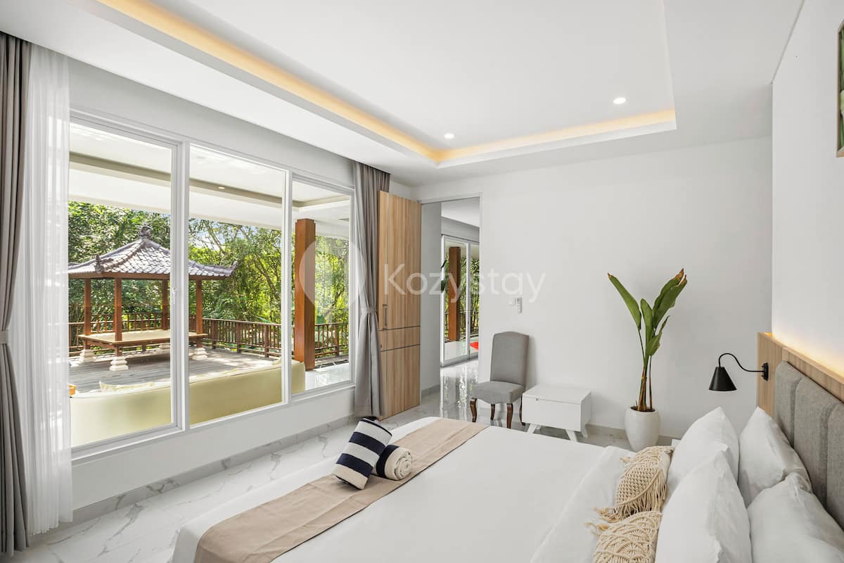 A modern hotel room with a large bed, a desk, a chair, and a view of a traditional pavilion through large windows.