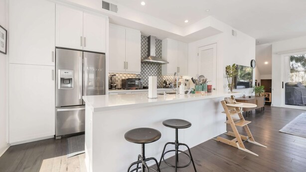 A modern kitchen with white cabinets, stainless steel appliances, a built-in island with stools, and a large window.