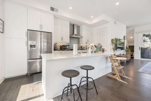A modern kitchen with white cabinets, stainless steel appliances, a built-in island with stools, and a large window.