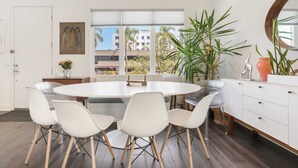 A modern kitchen with a round dining table, white chairs, a large potted plant, and a view of buildings outside.