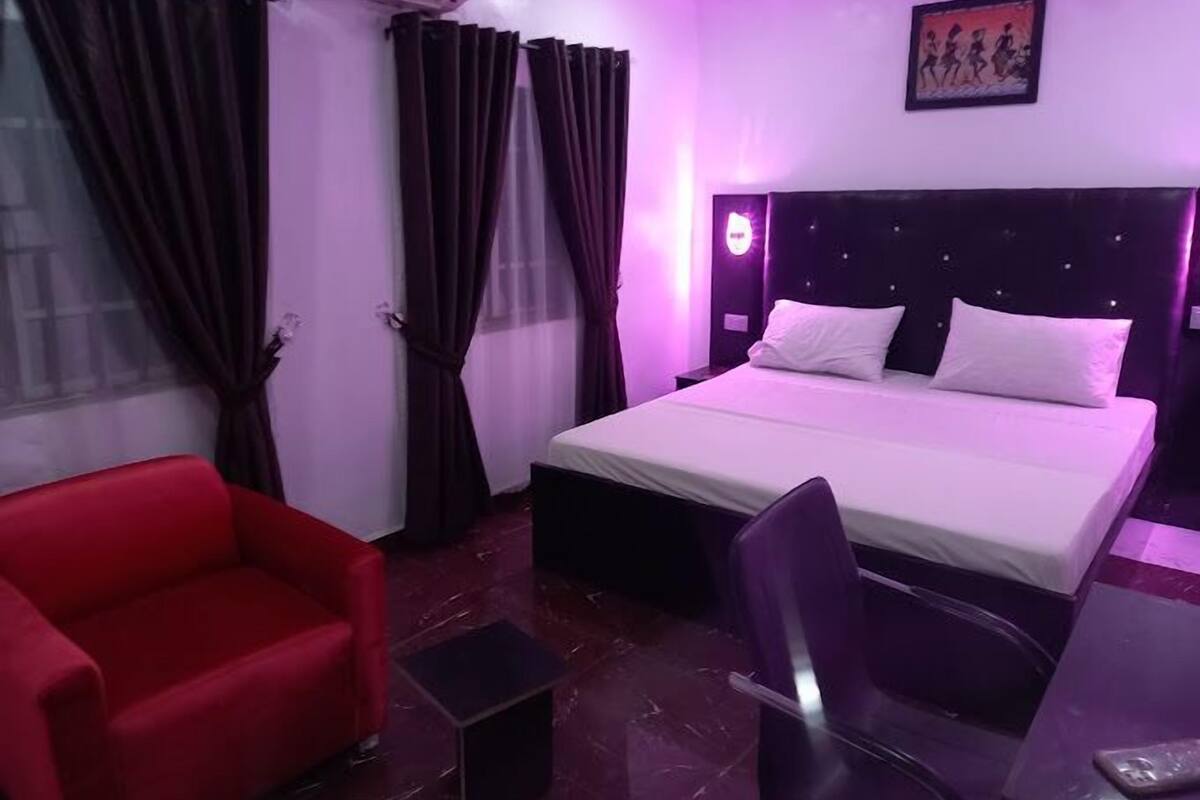 Ein Hotelzimmer mit einem Bett, einem roten Sessel, einem schwarzen Bürostuhl, einem kleinen Tisch und einer Wandklimaanlage.