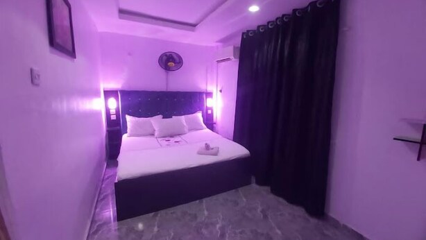 A bedroom with a bed, a nightstand, a wall-mounted clock, and a window with curtains. - De - Calmcity Guest House  (Ilorin)