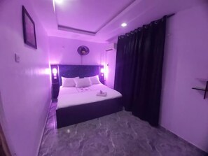 Un dormitorio con cama, mesita de noche, reloj en la pared y ventana con cortinas.