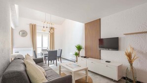 Ein modernes Wohnzimmer mit einem grauen Sofa, einem weißen Couchtisch, einem an der Wand montierten Fernseher und einem Essbereich mit Tisch und Stühlen.