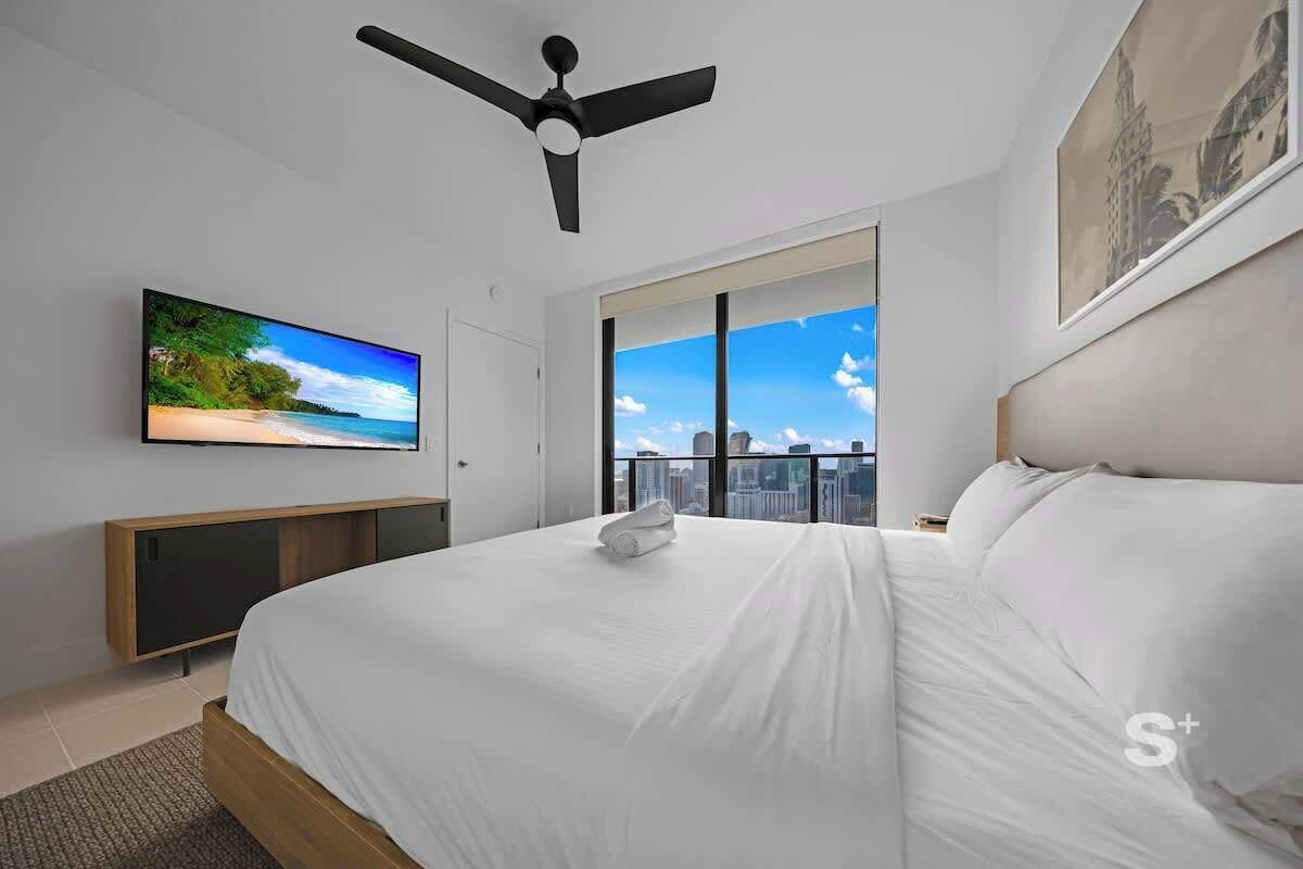 A modern hotel room with a large bed, a ceiling fan, a TV on the wall, and a city view through the window.