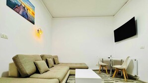 Una sala de estar con un sofá modular, una mesa de centro, una mesa de comedor con sillas, un televisor de pantalla plana y un cuadro en la pared.