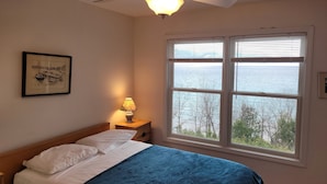Een slaapkamer met een bed, een nachtkastje met een lamp, een plafondventilator en een groot raam met uitzicht op zee.