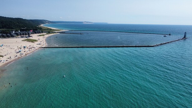 Luchtbeeld van een strand met een lange pier die in het water reikt, omringd door helder turquoise water en een zandstrand.