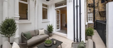 Un espacio exterior moderno con zona de estar, un sofá, sillas y una mesa de centro, rodeado de plantas en macetas y la fachada blanca de un edificio.