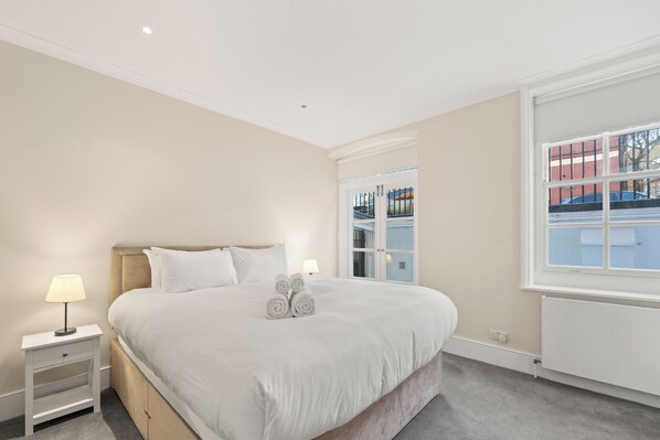 A bedroom with a large bed, bedside tables, and a window with a view of buildings. - Knightsbridge Luxury: 3BR/3BA, AC, Patio (London)