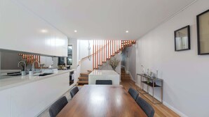 A modern kitchen with a wooden dining table and chairs, a staircase with orange railings, and a small side table with decorative items.