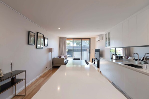 A modern kitchen with a large island, a dining area with a sofa, and a view through a sliding glass door to the outdoors.