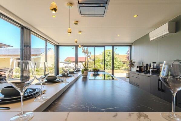 A modern kitchen with a long countertop, overhead lighting, and large windows offering a view of the outdoors.