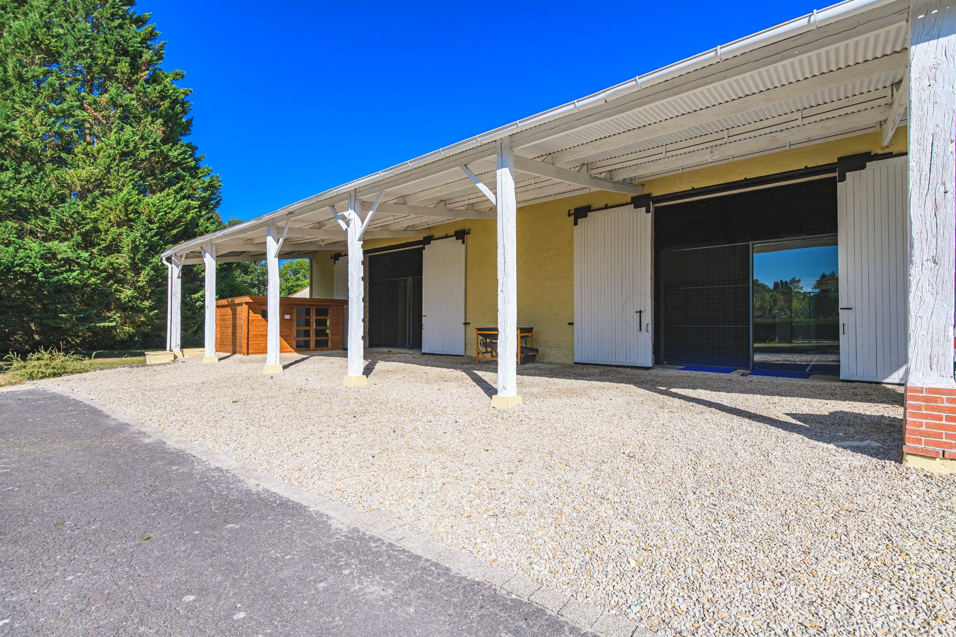 A building with a covered outdoor area, a gravel driveway, and a wooden structure to the left.